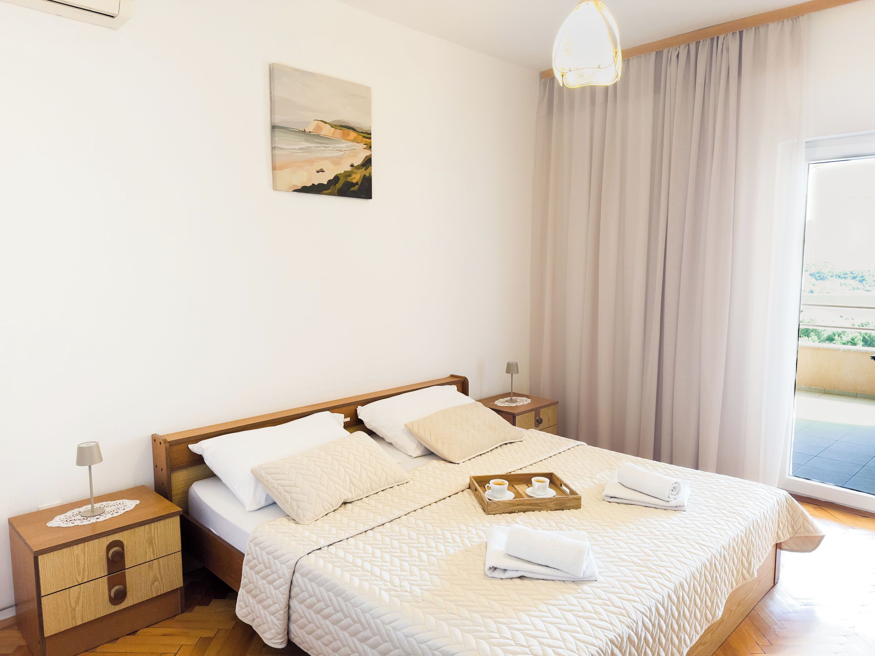 A neatly made bed with a quilted cover, a bedside table with a lamp, a nightstand with a drawer, and a painting on the wall.