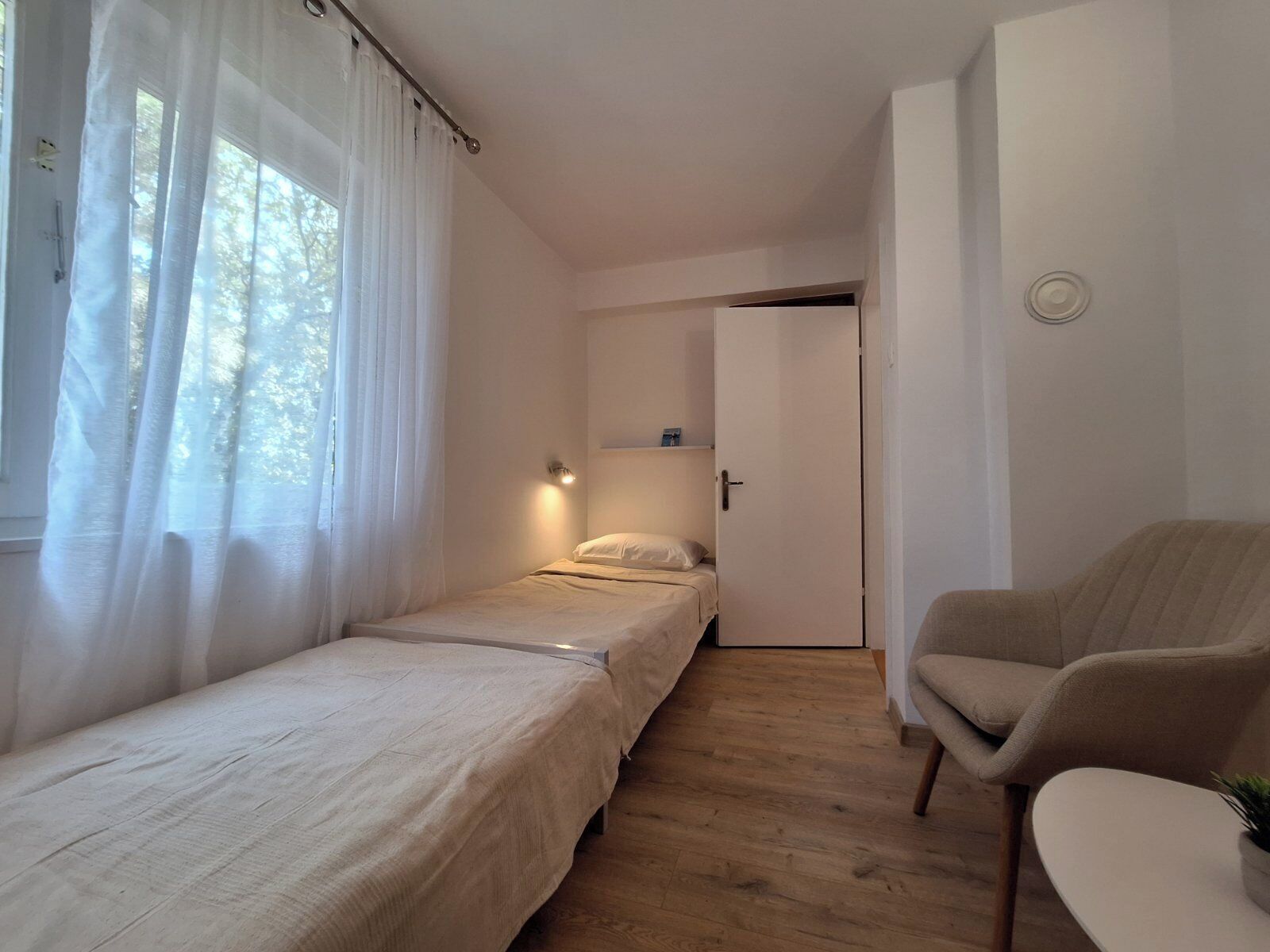 A small, well-lit bedroom with a single bed, a chair, and a door leading to another room.