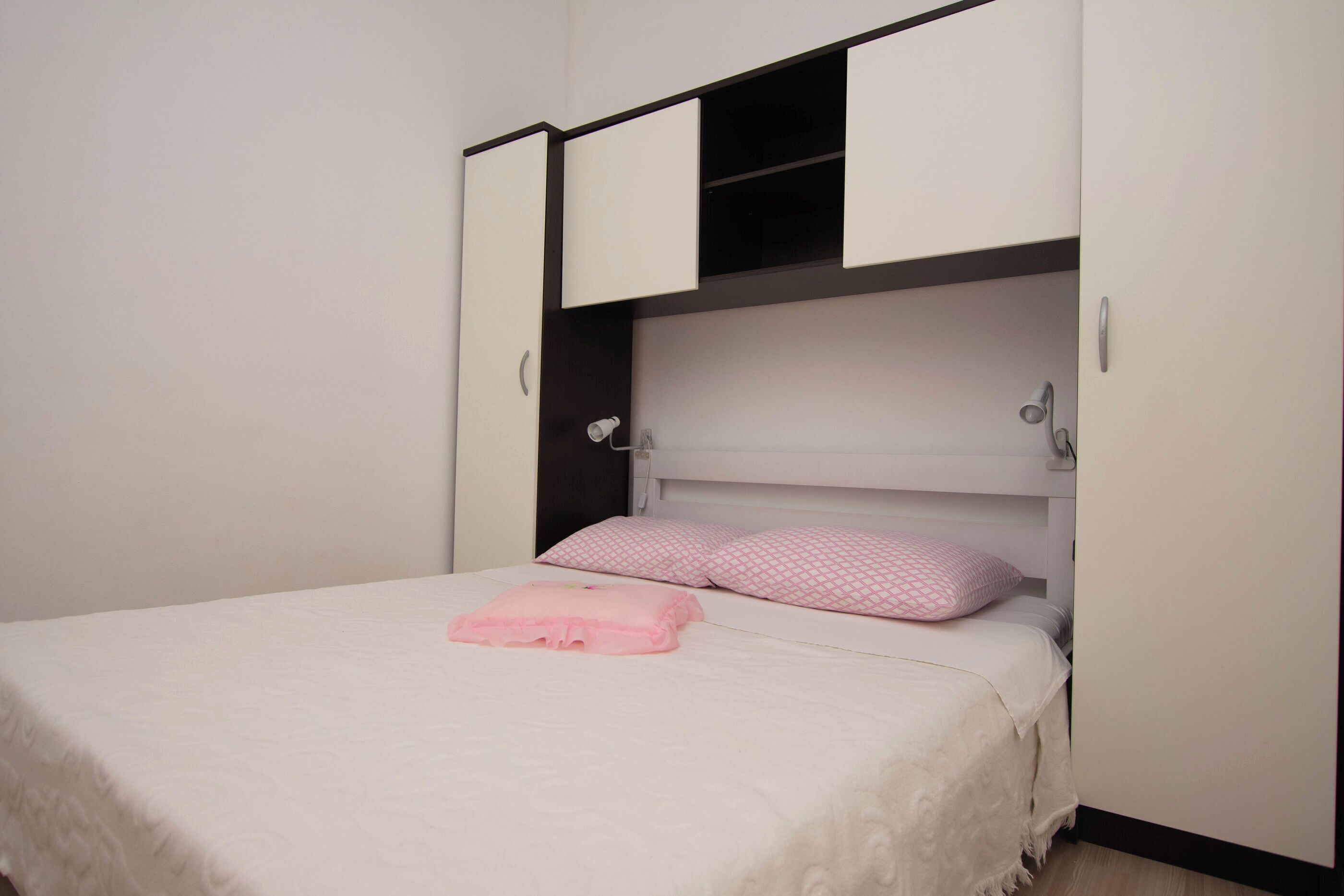 Un lit avec une couvre-lit blanche et un oreiller rose, une tête de lit blanche et une armoire dont les portes sont ouvertes.
