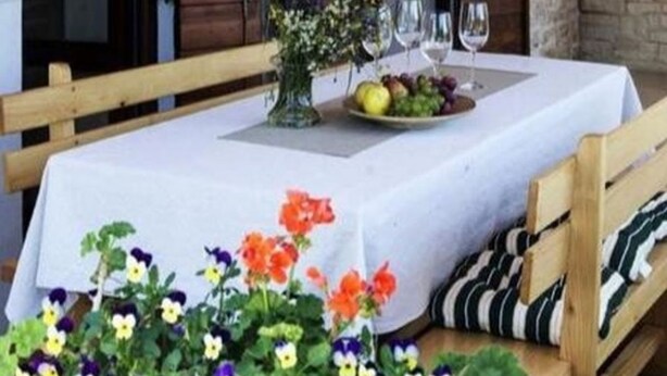 Un espacio exterior para comer con una mesa lista para dos personas, un jarrón de flores y un frutero.