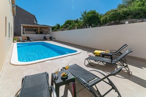 En swimmingpool med liggestole og et bord, omgivet af en fliseterrasse og en lav stenvæg.