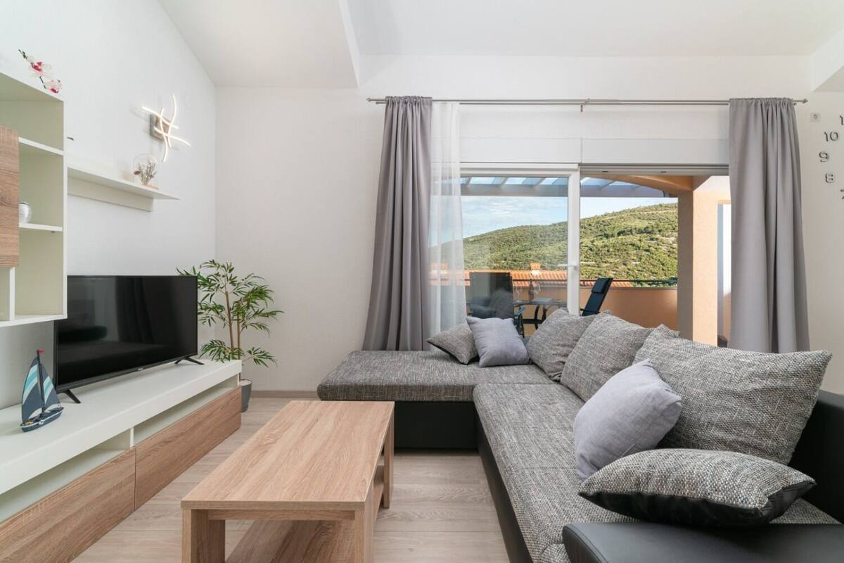 A modern living room with a sectional sofa, a flat-screen TV, a wooden coffee table, and a large window with curtains.