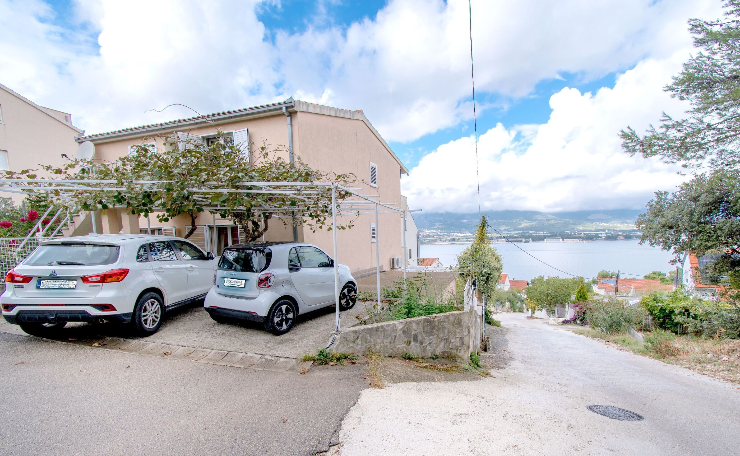 A residential street with two parked cars, a building with a sloped roof, and a view of a lake and mountains in the distance.