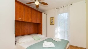 A bedroom with a bed, wooden wardrobe, ceiling fan, window with curtains, and a framed picture on the wall. - Apartment Ivan (Solta)
