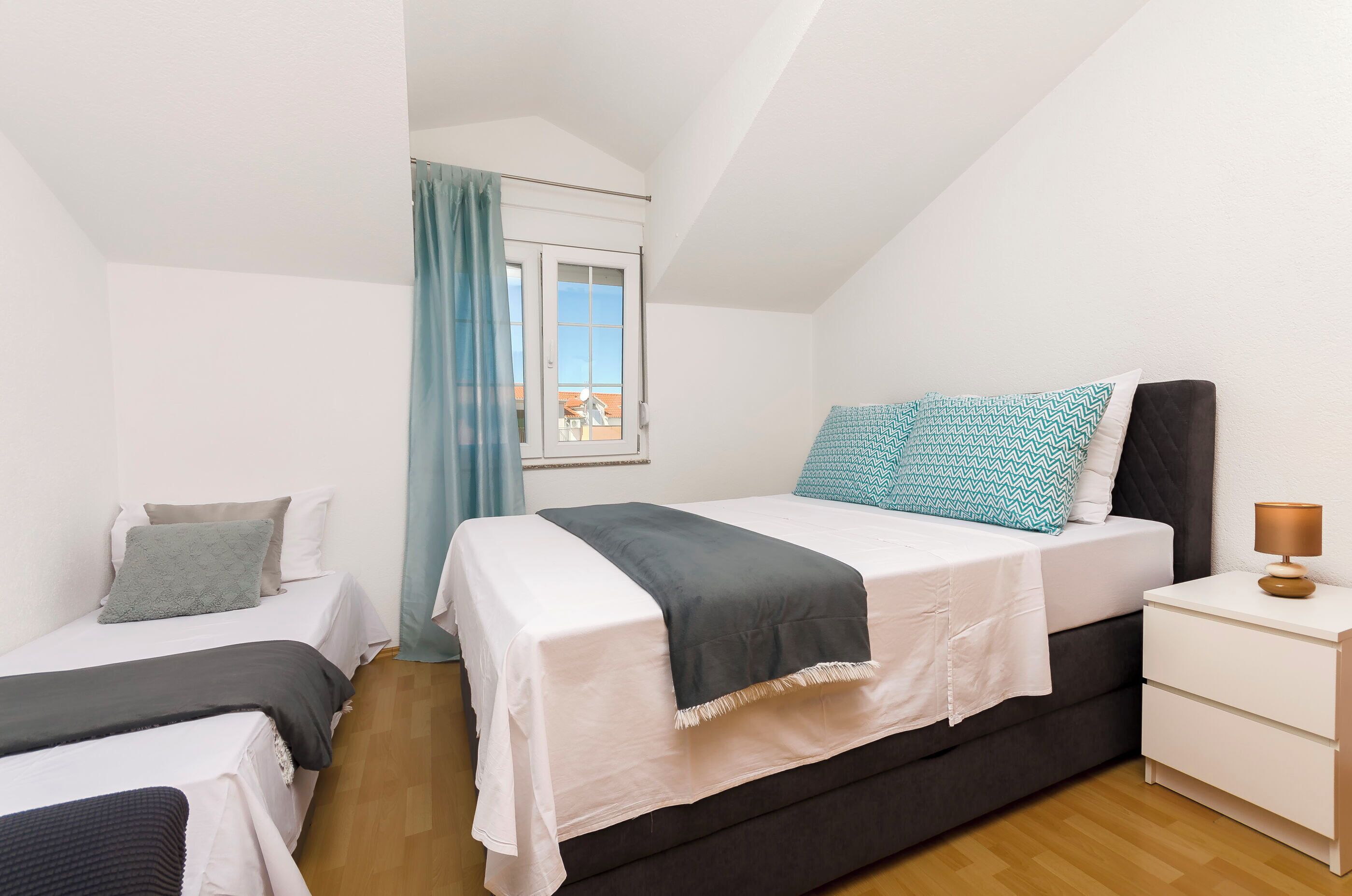 A small, well-lit bedroom with two beds, a window with curtains, a nightstand with a lamp, and a view of the outside.