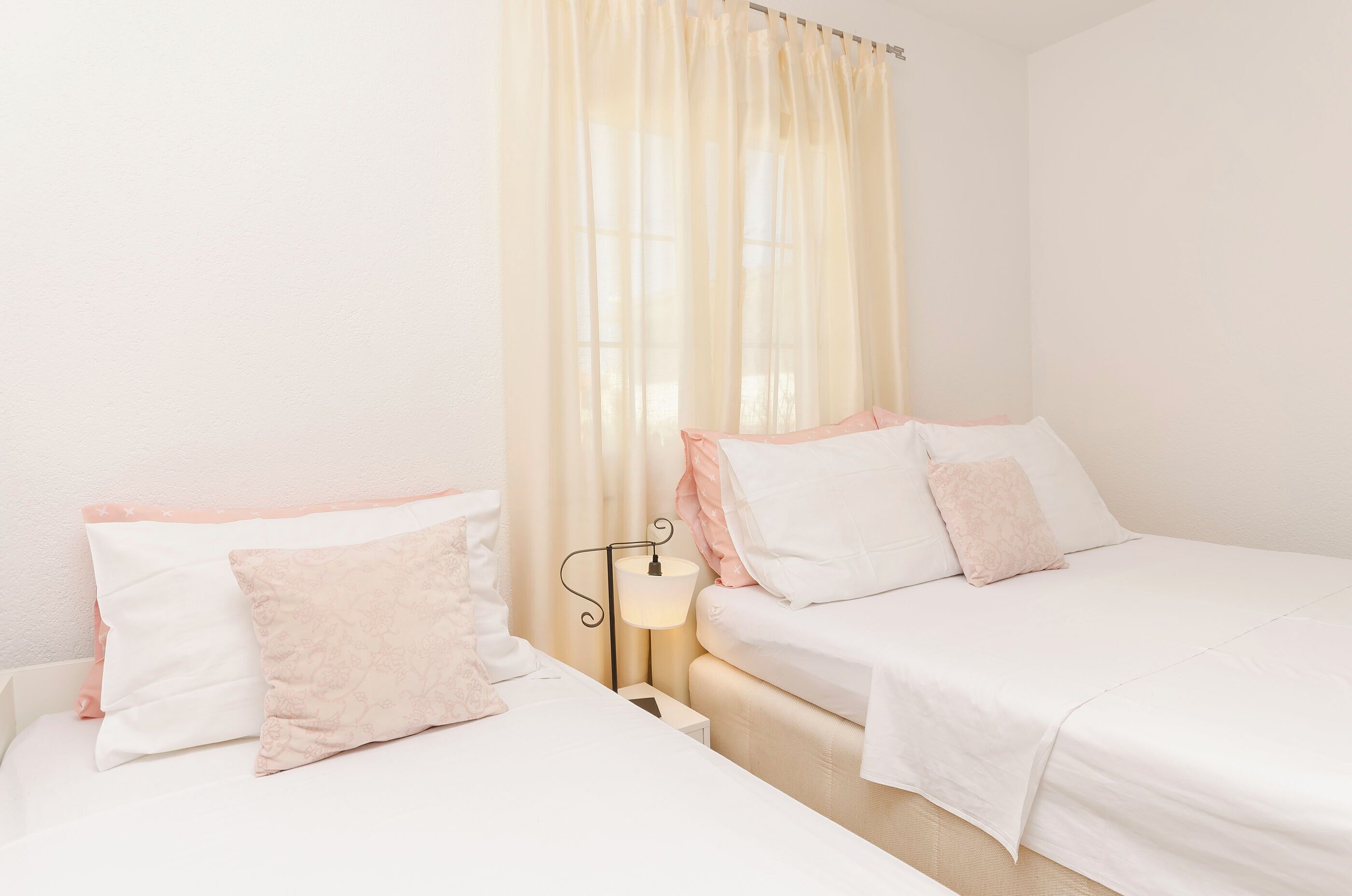 A bedroom with two beds, a bedside table, a lamp, and sheer curtains.
