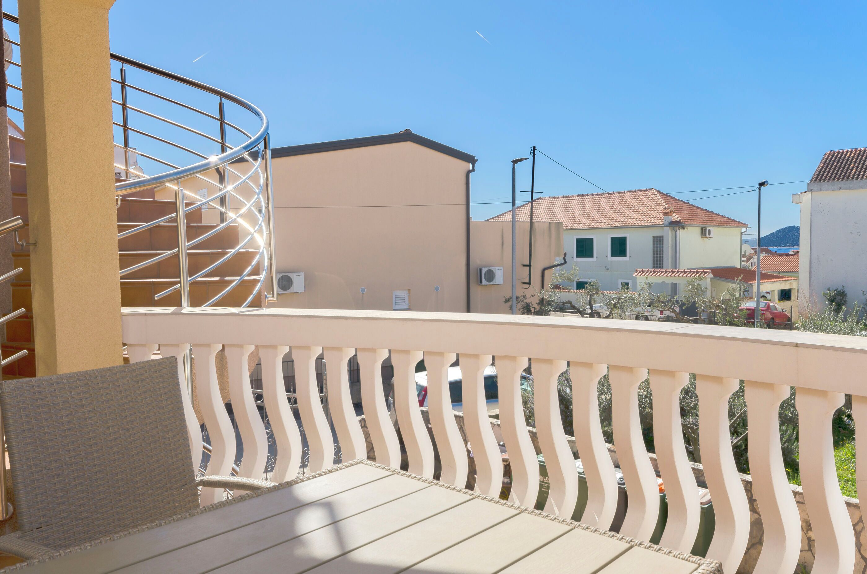 Ein Balkon mit Metallgeländer, einem weißen Sitzkissen und Blick auf eine Wohngegend mit Häusern und parkenden Autos.