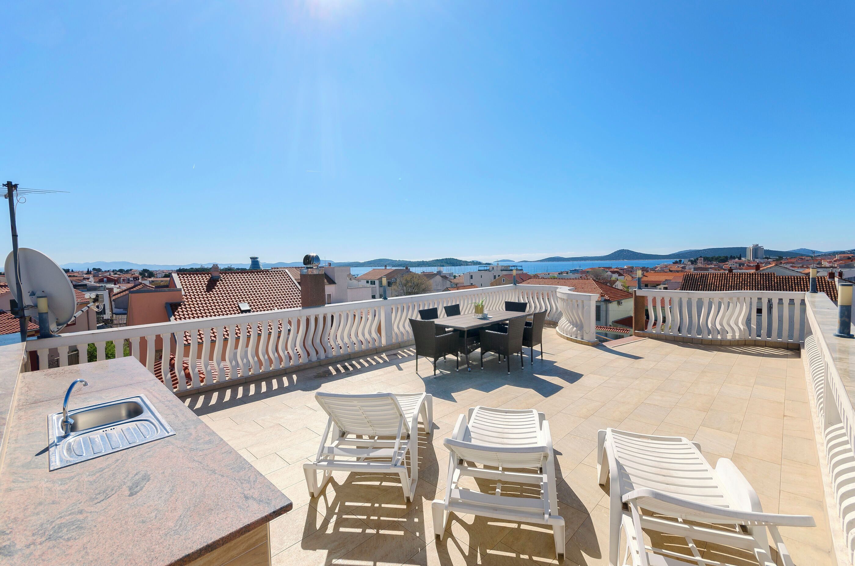 A rooftop terrace with a sink, white chairs, a table, and a view of the city and hills.