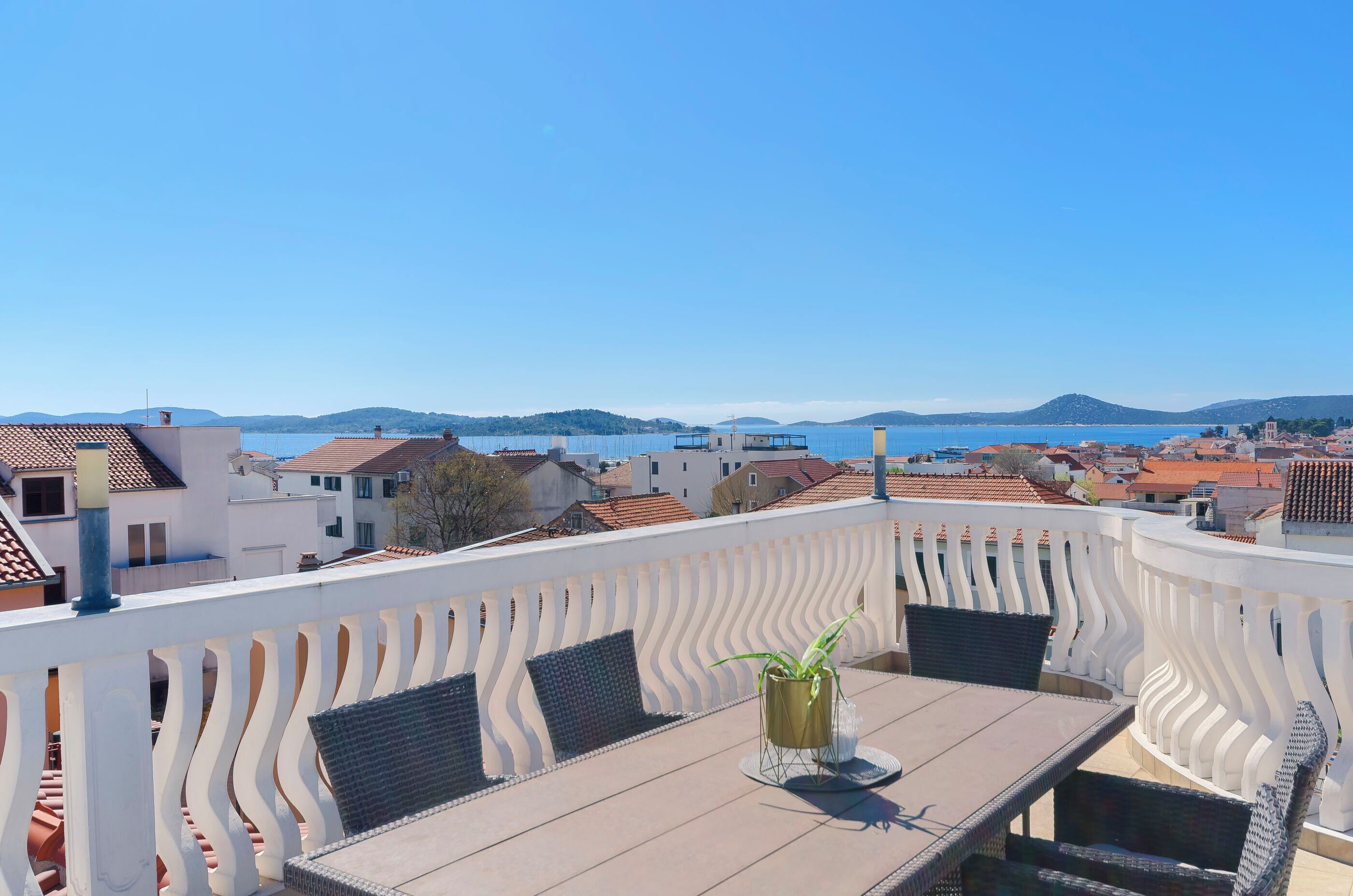 A terrace with a table and chairs, a potted plant, and a view of a coastal town and hills.