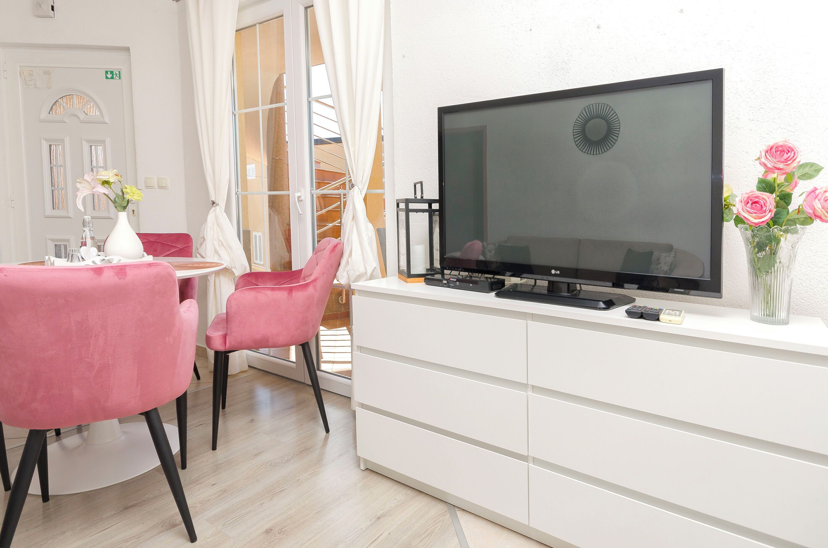Ein modernes Zimmer mit einem Flachbildfernseher auf einer weißen Kommode, rosa Stühlen, einer weißen Vase mit Blumen und einer sichtbaren Treppe durch eine offene Tür.
