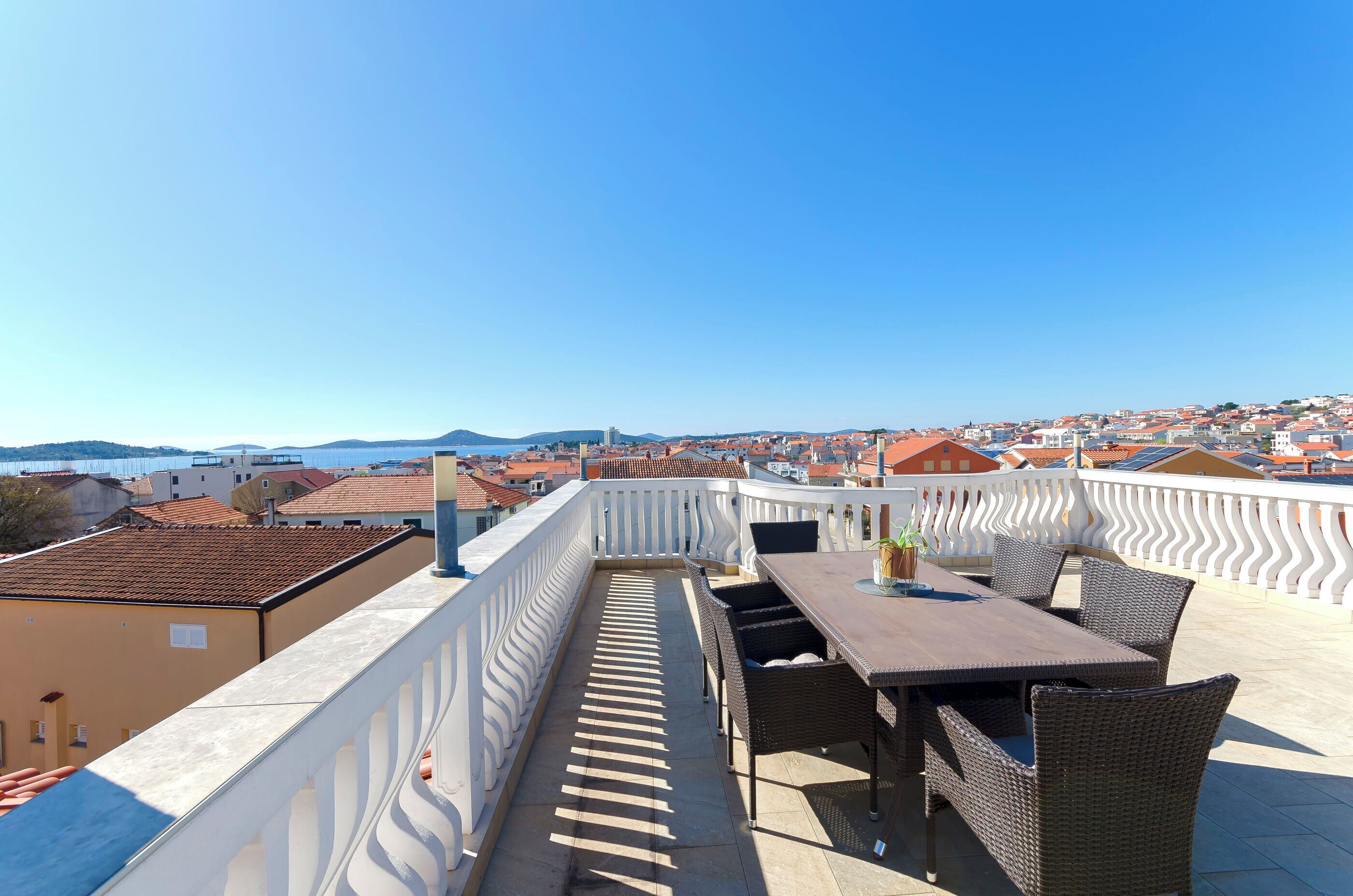 Una terrazza con un tavolo e sedie, affacciata su una cittadina costiera con edifici dai tetti rossi.