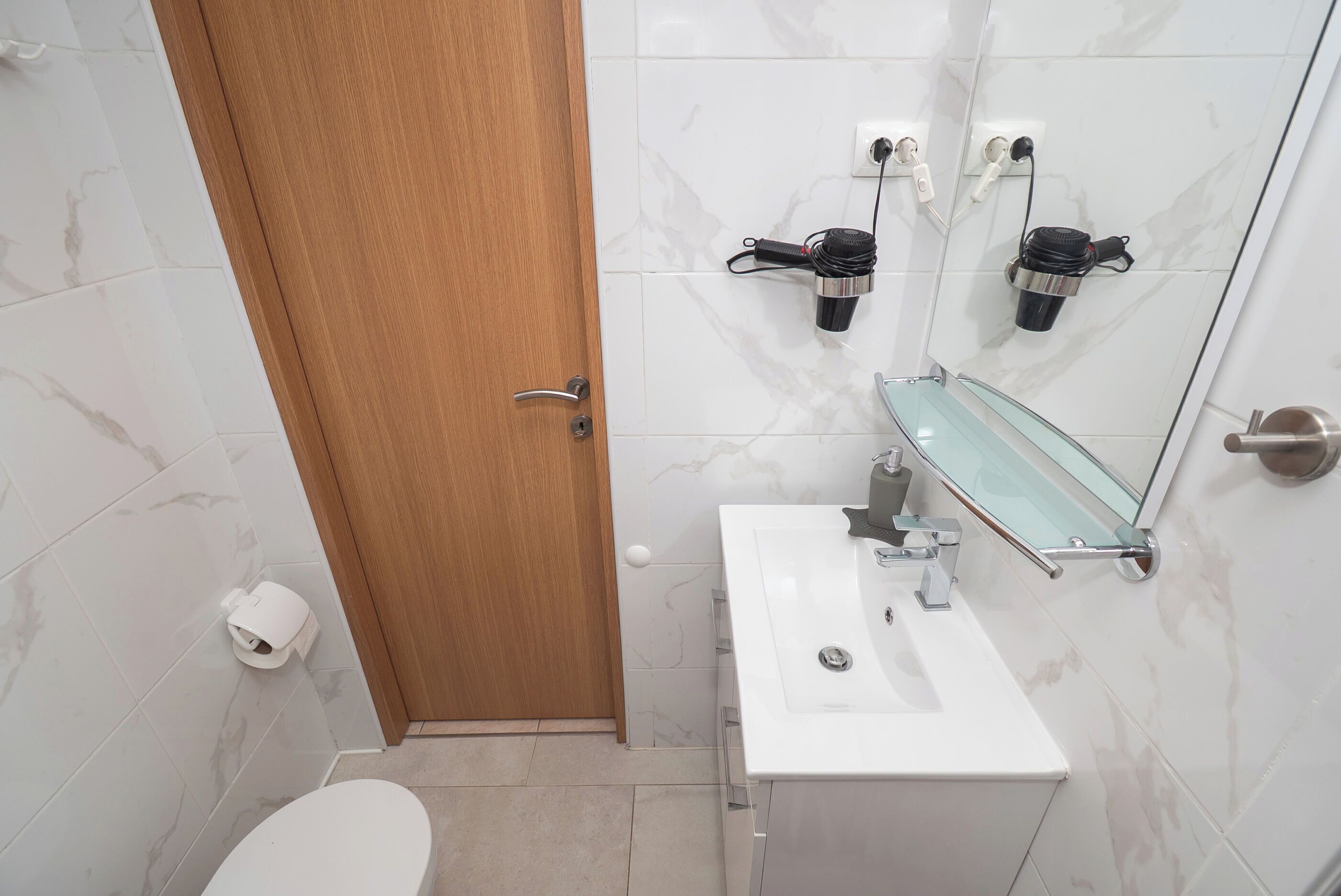A bathroom with a white sink, a glass shelf, a wooden door, and a toilet.
