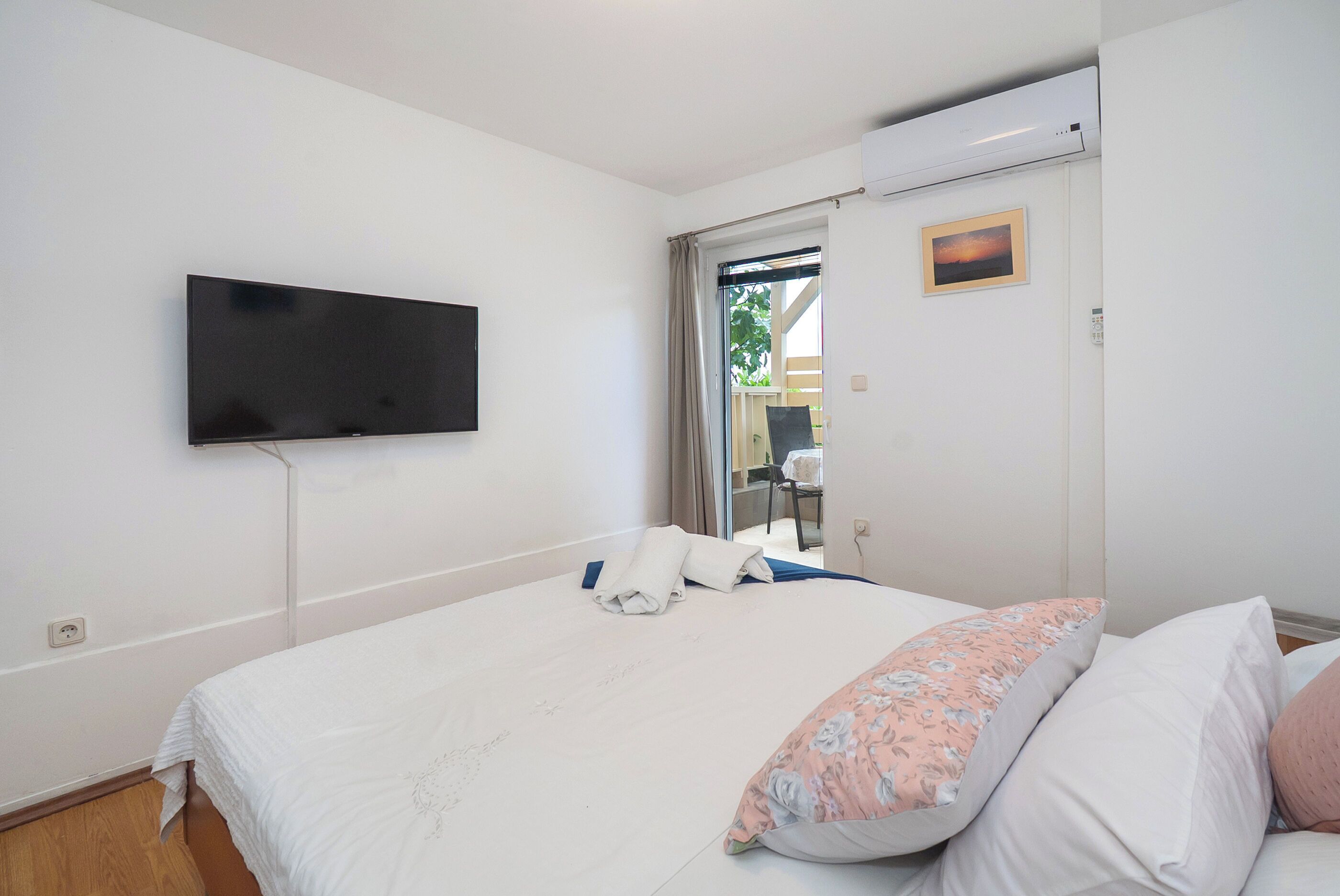 A bedroom with a bed, pillows, a flat-screen TV mounted on the wall, and a sliding door leading to a balcony.