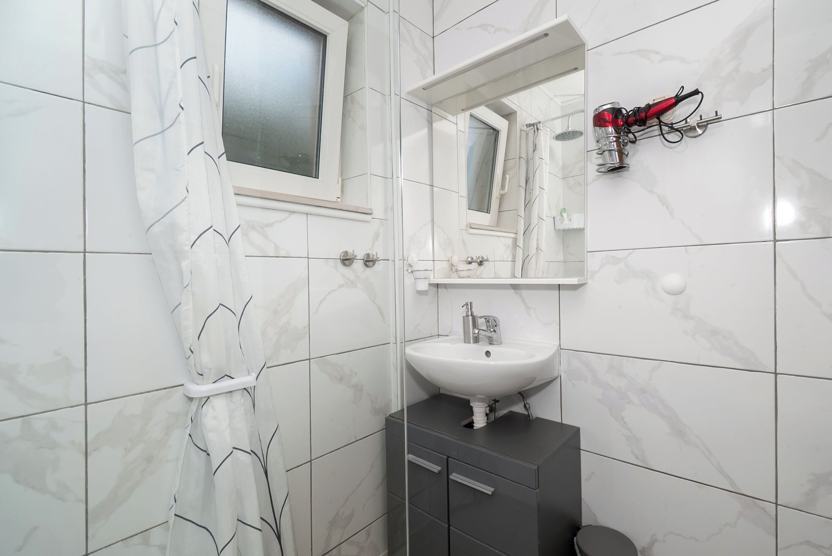 A bathroom with white marble tiles, a white sink with a mirror above it, a window, and a red hairdryer on the wall.