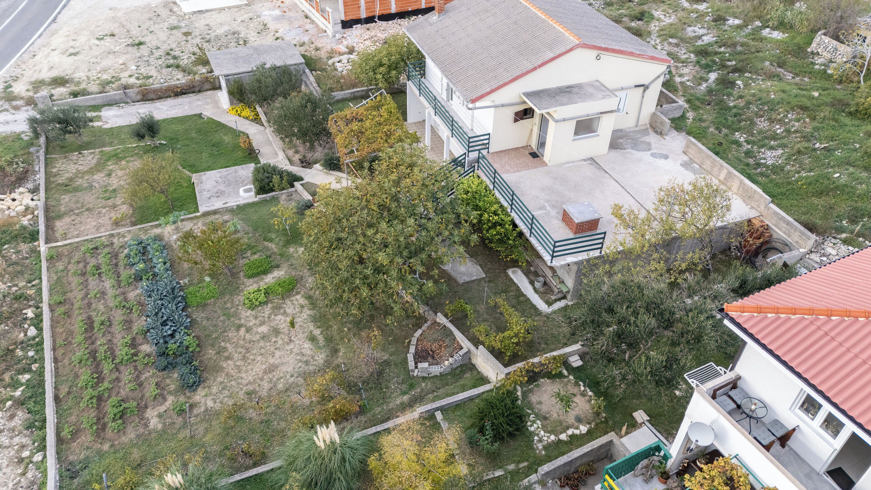 An aerial view of a residential area with a white house featuring a balcony, a garden with various plants, and a red-roofed building to the right.