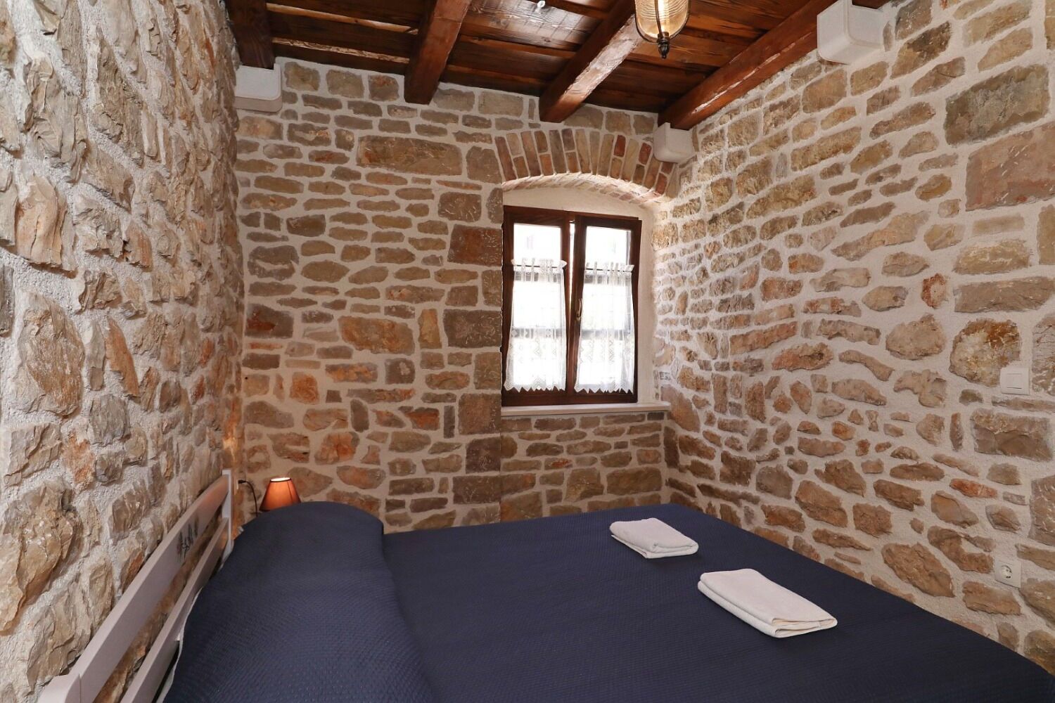 A small, stone-walled room with a single bed, a window with curtains, and a wooden ceiling.