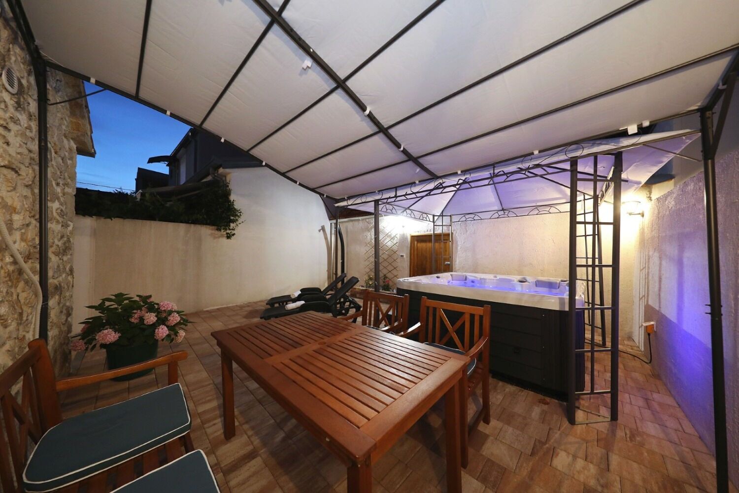 Un porche cubierto con una mesa y sillas de madera, un jacuzzi y un muro de piedra.