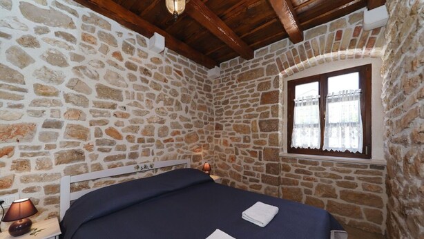 Une chambre avec un mur en pierre, un lit recouvert d’une couvre-lit bleu foncé, une lampe de chevet et une fenêtre ornée de rideaux en dentelle.