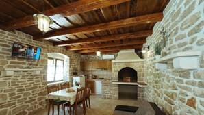A rustic kitchen with stone walls, wooden beams, a stone oven, and a dining area with a table and chairs. - House Cirrus (Sukosan)