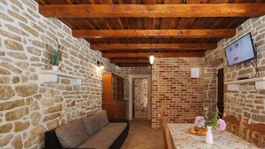 A cozy room with a stone wall, wooden beams, a sofa, a table with a floral tablecloth, and a television. - House Cirrus (Sukosan)