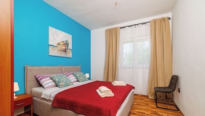 A bedroom with a bed, a chair, a nightstand, a window with curtains, and a painting on the wall. - Apartments Domagoj (Karlobag)