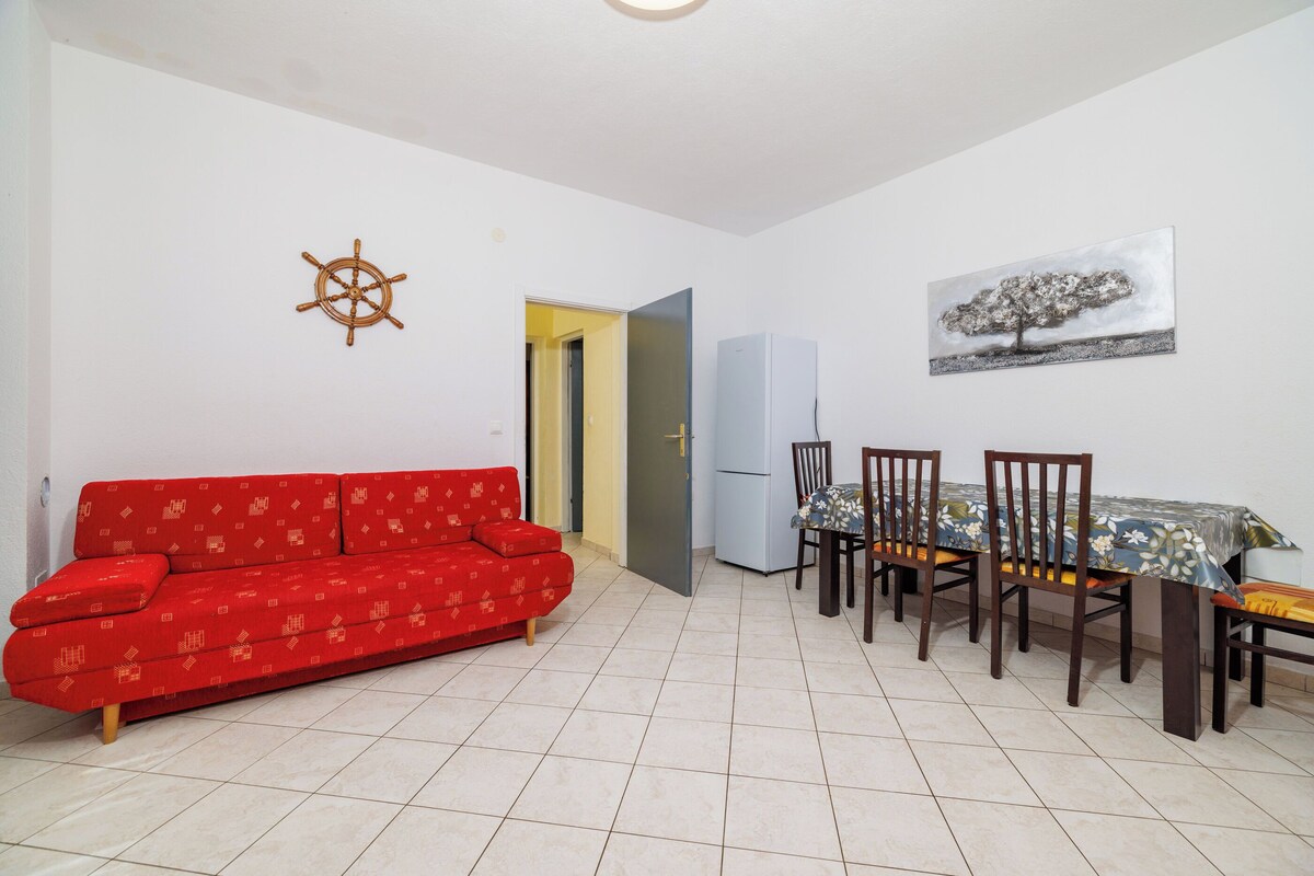 Ein Wohnzimmer mit einem roten Sofa, einem Esstisch mit Stühlen, einem Kühlschrank sowie einem Wandbild mit Schiffsrad und Baum.