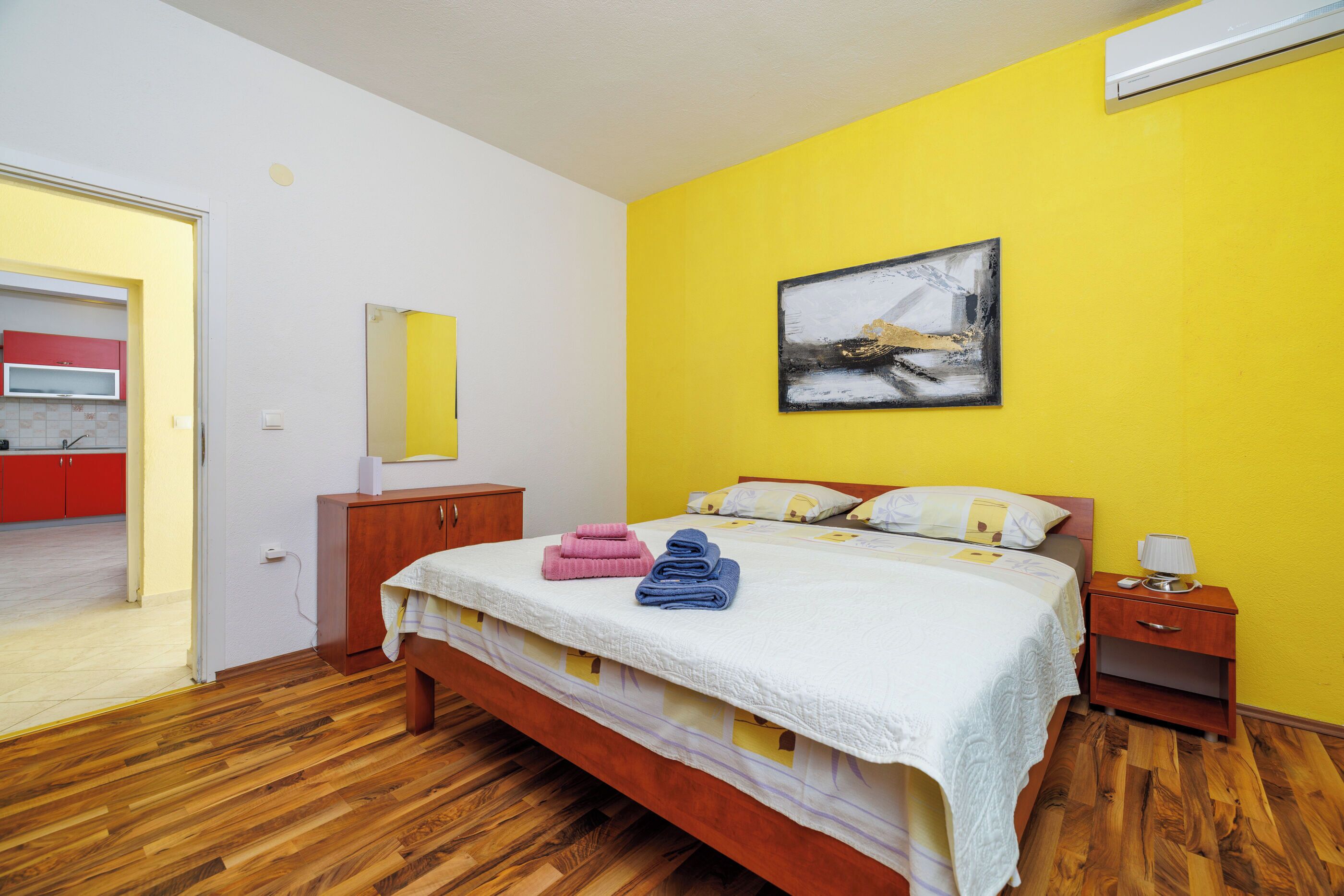 Un dormitorio con cama, suelo de madera, una mesita de noche y un cuadro en la pared, con vistas a una zona de cocina.