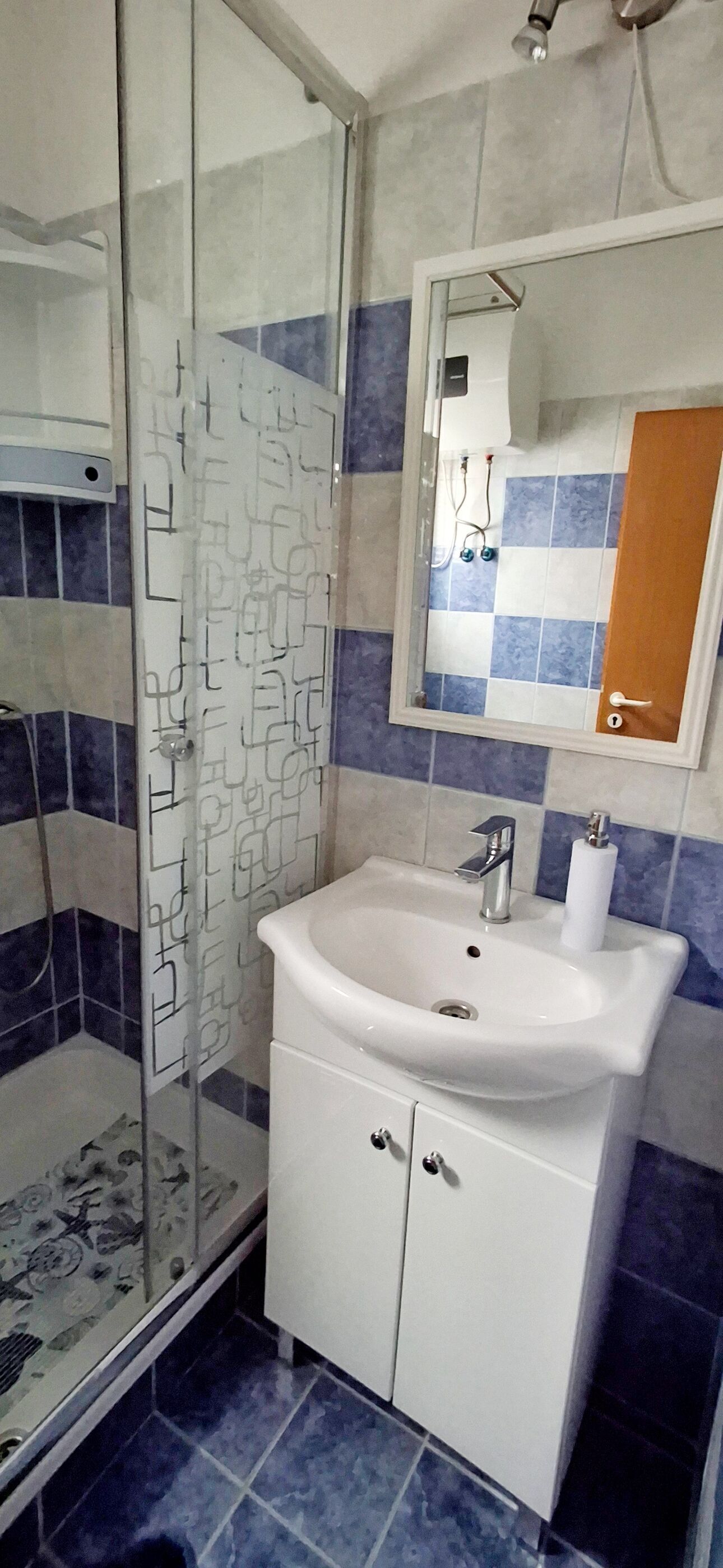 Un baño con cabina de ducha de vidrio, un lavamanos blanco con espejo encima y paredes con azulejos azules y blancos.