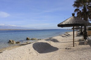 Een strand met een rieten parasol, kiezels en een palmboom.