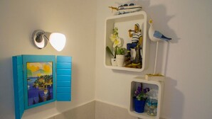 Un baño con espejo de marco azul, luz empotrada en la pared y estantes con adornos como una maqueta de barco, flores y una figurita de pájaro.