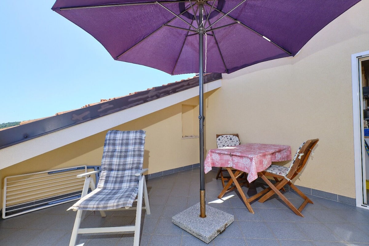 Un balcón con una sombrilla morada, una silla lounge blanca, una mesa de madera con mantel de flores y dos sillas de madera.