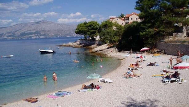 Une plage où l’on voit des gens se prélasser au soleil et nager, des bateaux sur l’eau et des bâtiments en arrière-plan.
