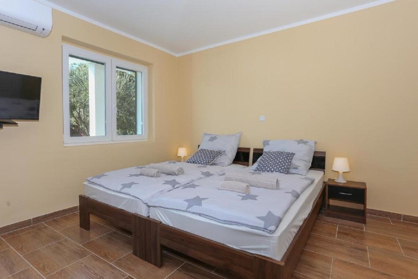 A bedroom with a bed, two bedside tables, a TV mounted on the wall, and a window with a view of trees.