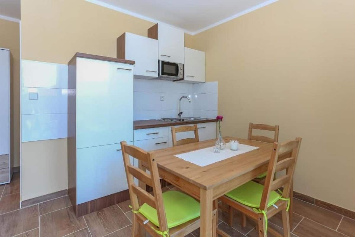 Een kleine keuken met witte kastjes, een magnetron en een spoelbak. Er staat een houten eettafel met vier stoelen, elk voorzien van een groene kussentje.