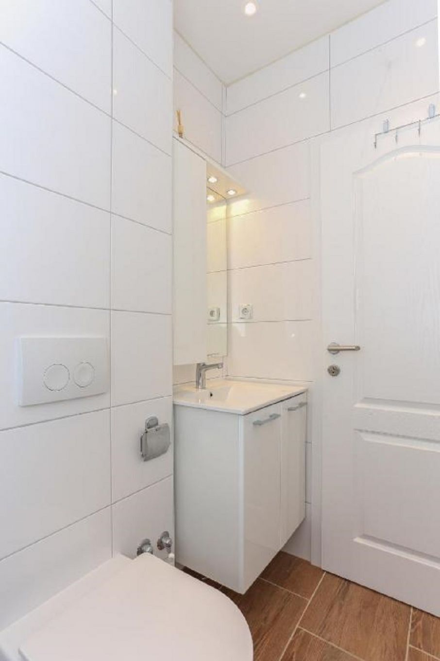 A modern bathroom with white tiles, a toilet, a sink with a mirror, and a door.