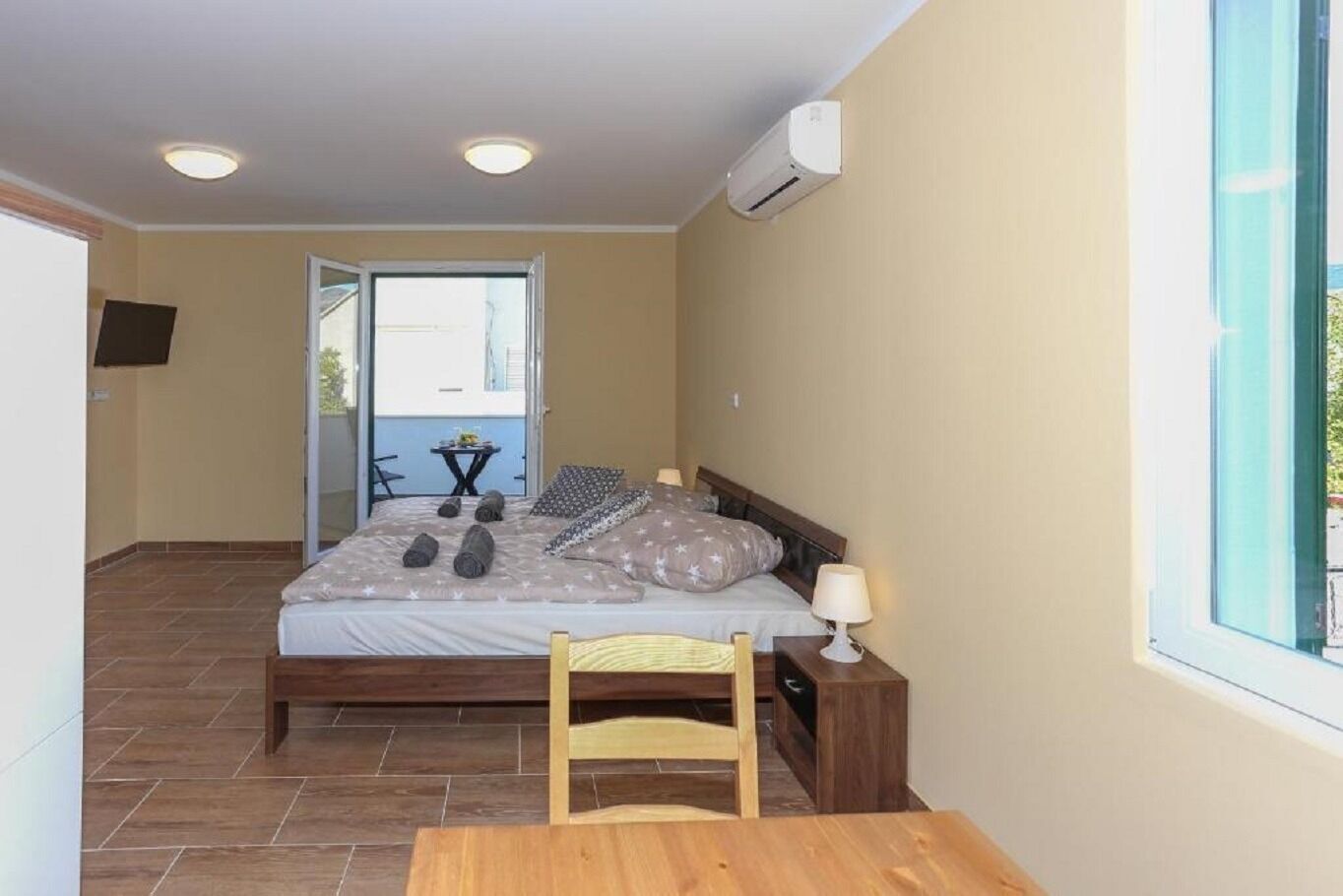 A bedroom with a bed, bedside table, lamp, and a view of the outside.