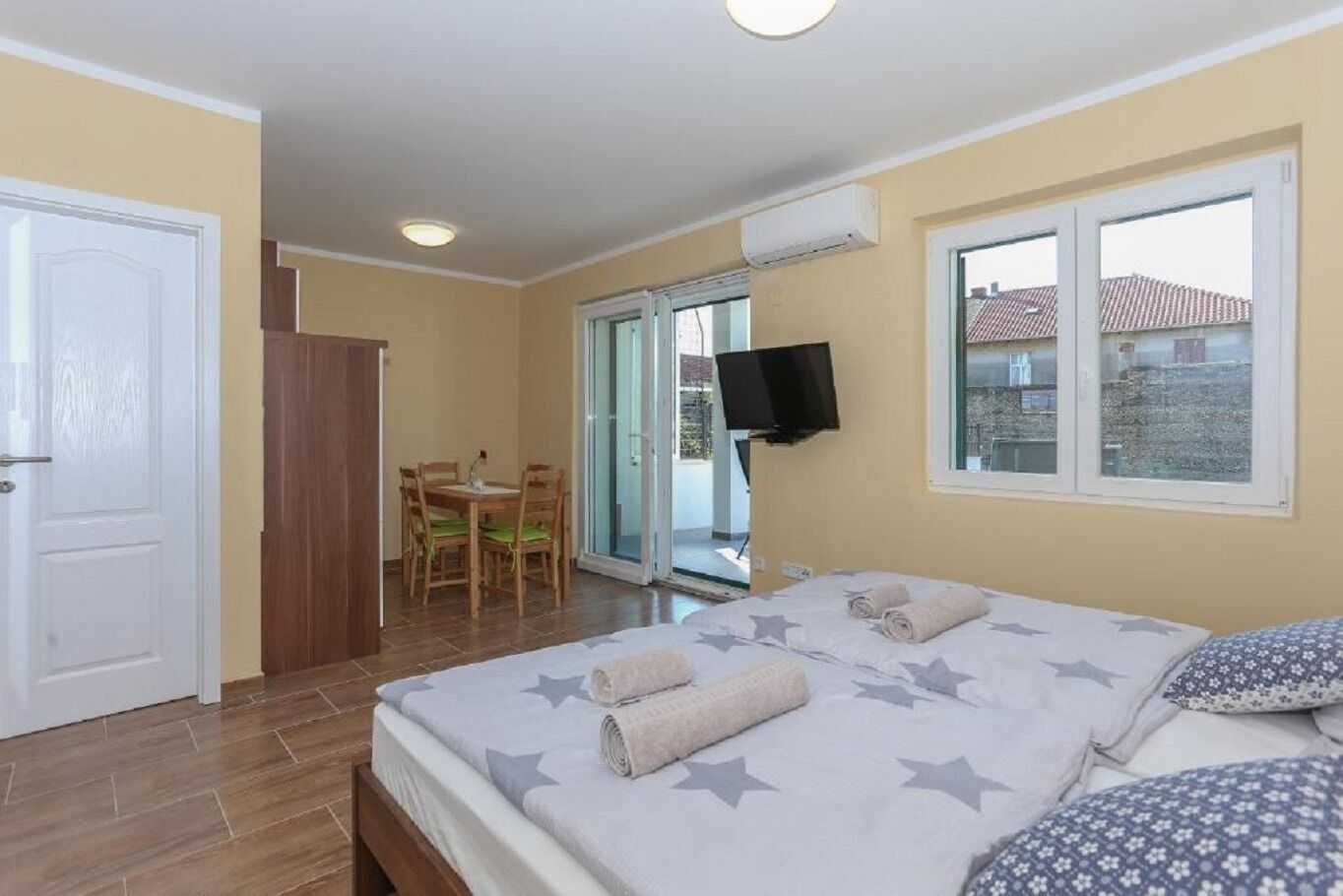 A hotel room with a bed, a TV mounted on the wall, a dining table with chairs, and a sliding glass door leading to an outdoor area.