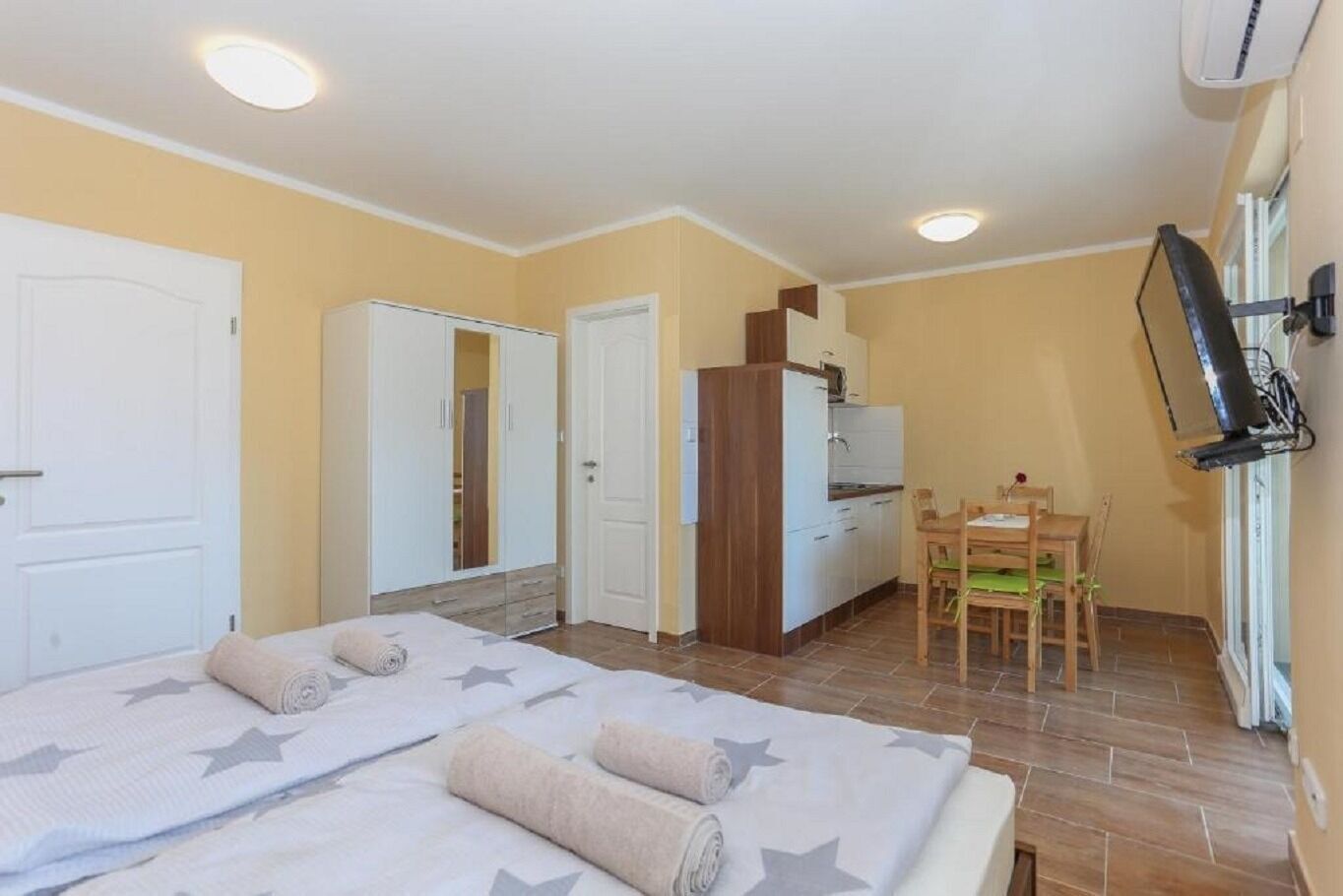Quarto com duas camas, uma kitchenette, zona de refeições e televisão na parede.