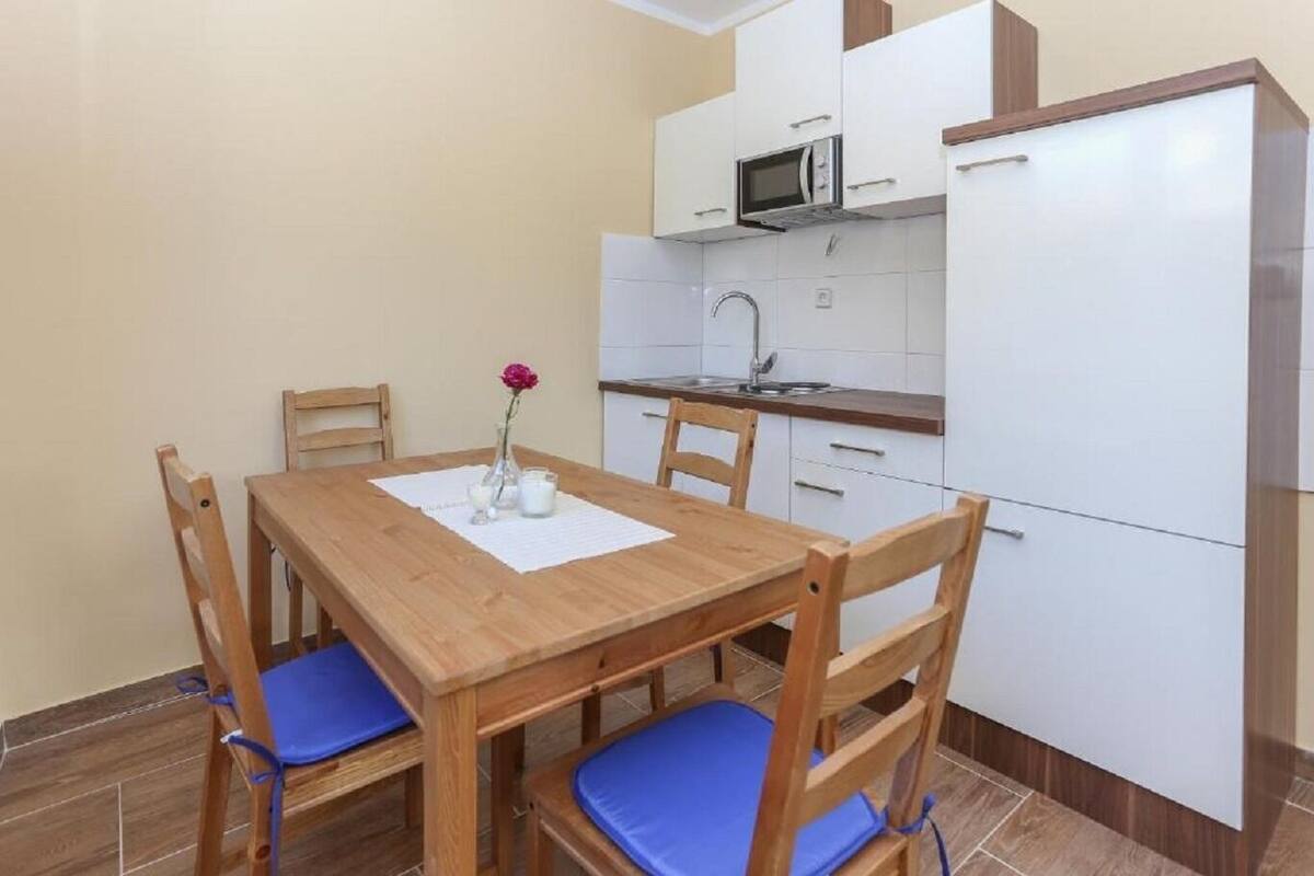 Een compacte keuken met witte kastjes, een magnetron en een koelkast. Er staat een houten eettafel met blauwe stoelen met kussentjes en een vaas met een bloem op de tafel.