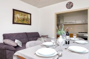 A dining area with a table set for four, a sofa, a painting, and a kitchen visible in the background. - Apartment Denko by the sea (Tisno)