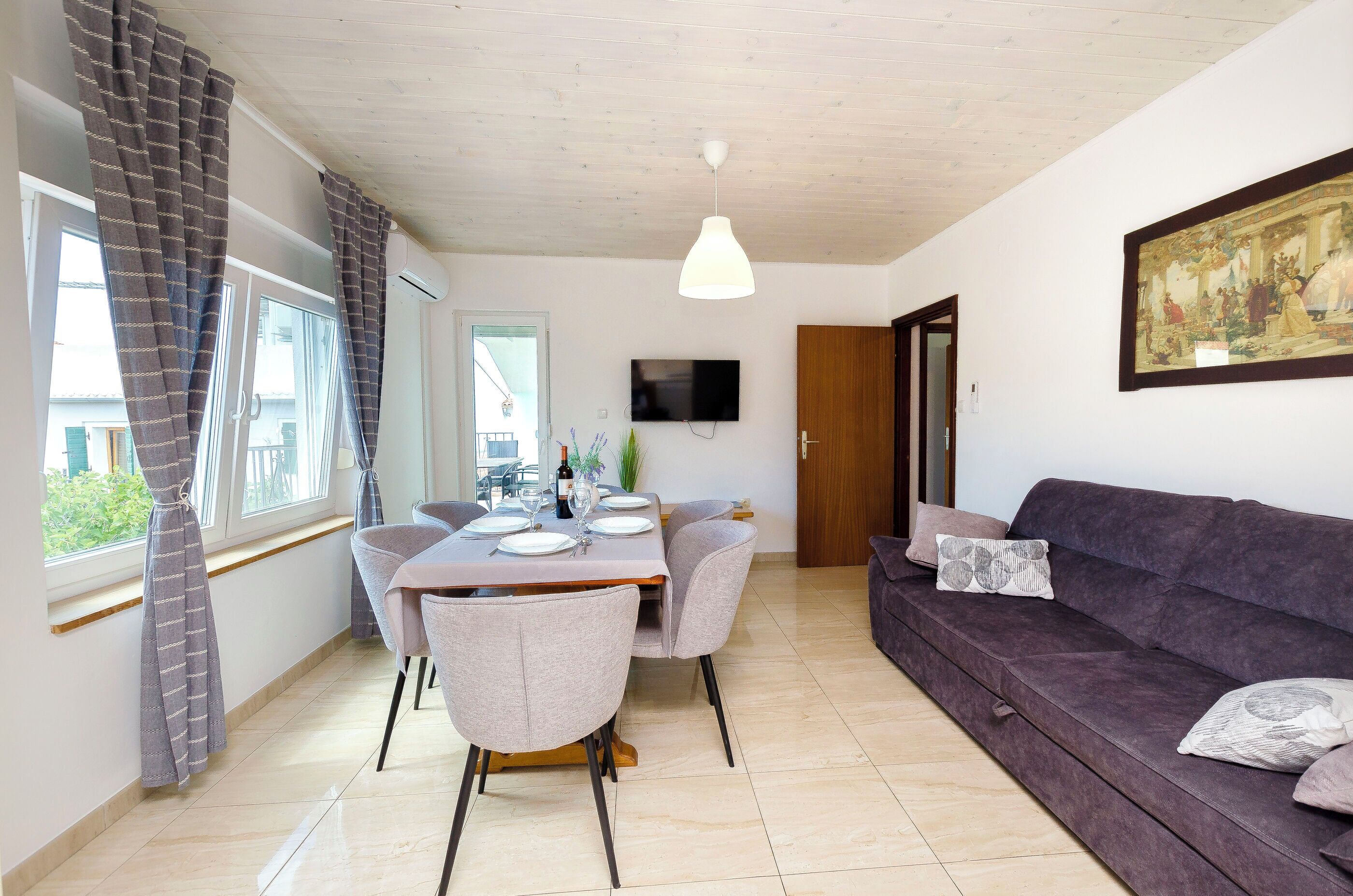 Een eetgedeelte met een tafel voor vier personen, een bank, een aan de muur bevestigde televisie en een deur die naar een andere kamer leidt.