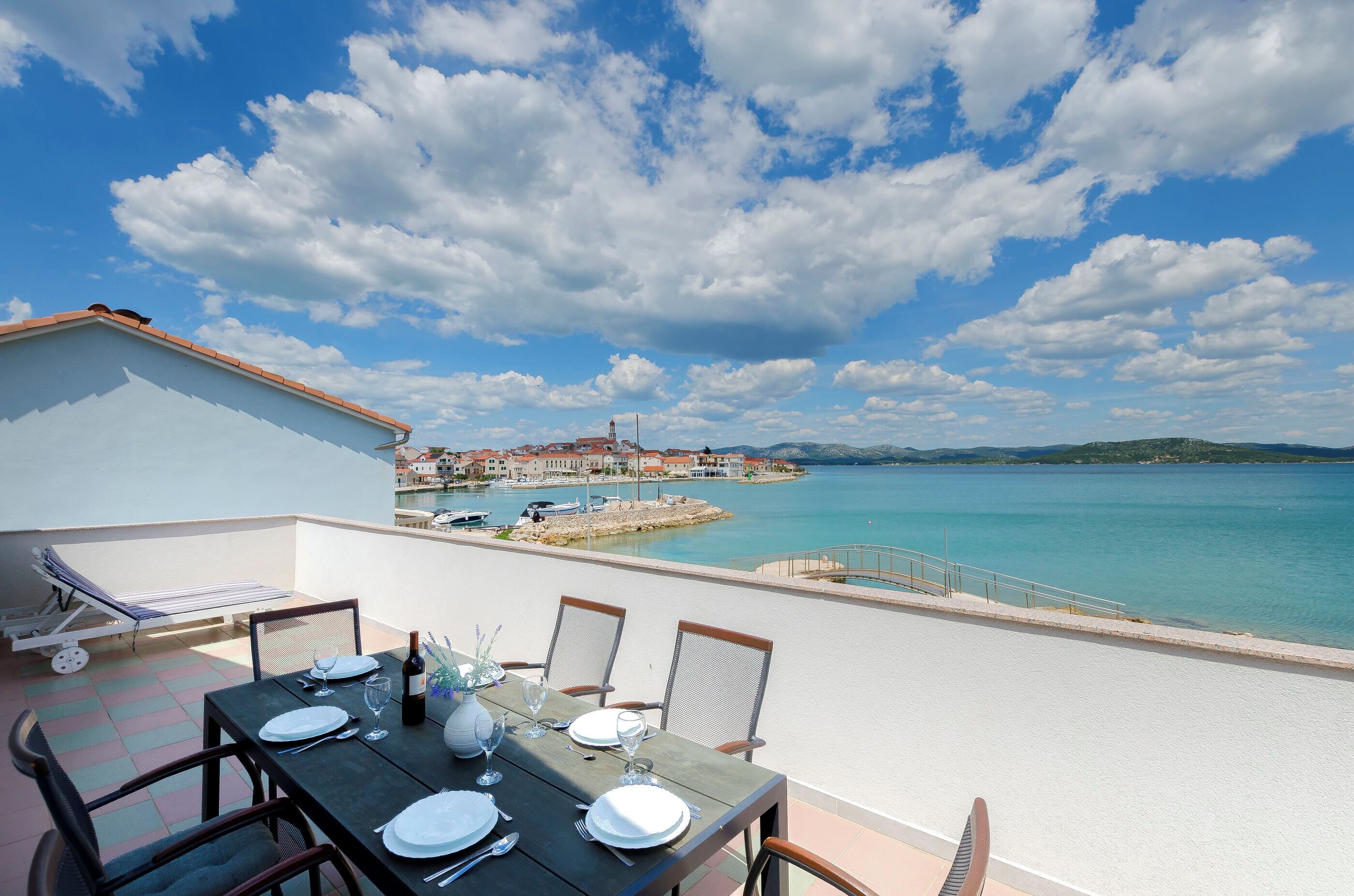 A dining table set with plates, glasses, and cutlery on a balcony overlooking a coastal town with a clear sky.