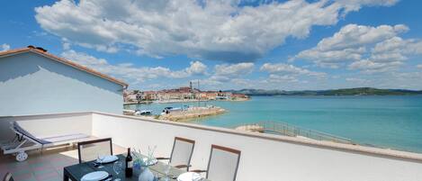 A dining table set with plates, glasses, and cutlery on a balcony overlooking a coastal town with a clear sky.