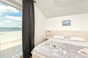 A bedroom with a bed, a nightstand, a picture on the wall, and a view of the sea through a window. - Apartment Denko by the sea (Tisno)