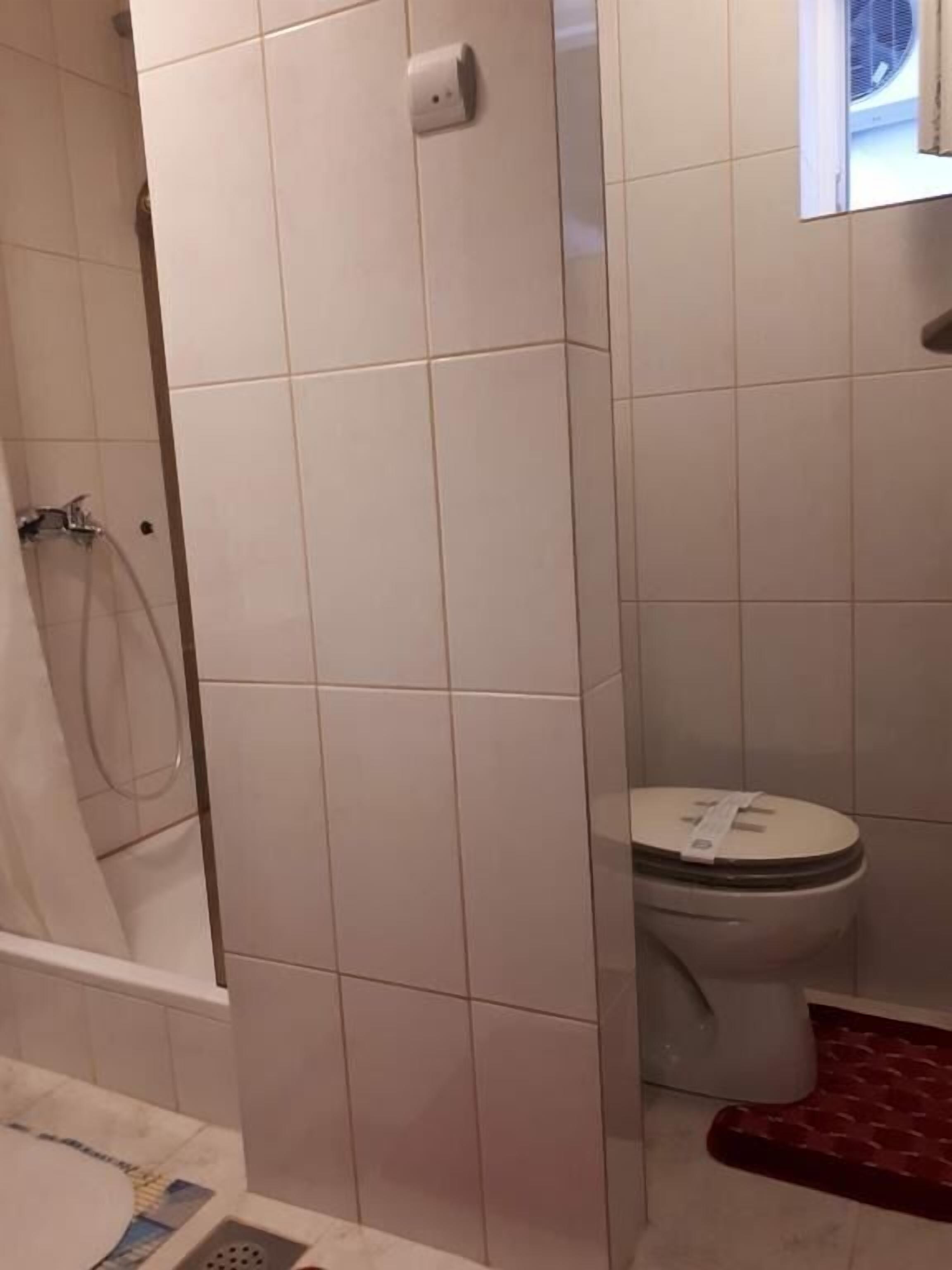 Un bagno con piastrelle bianche, un water, un angolo doccia e un tappetino rosso.