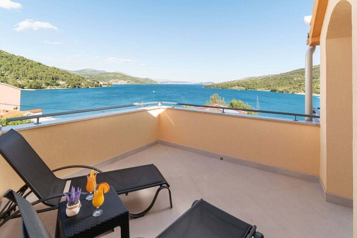 En balkon med to sorte liggestole, et lille bord med en potteplante og drikkevarer, samt udsigt over et blåt hav og grønne bakker.