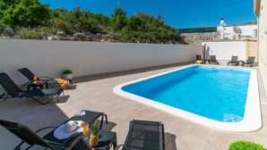 A rectangular swimming pool with a white edge, surrounded by beige tiles. There are black lounge chairs and a table with a pineapple and a drink on it. - Apartments Lux 2 (Marina)