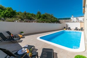 A rectangular swimming pool with a white edge, surrounded by beige tiles. There are black lounge chairs and a table with a pineapple and a drink on it. - Apartments Lux 2 (Marina)