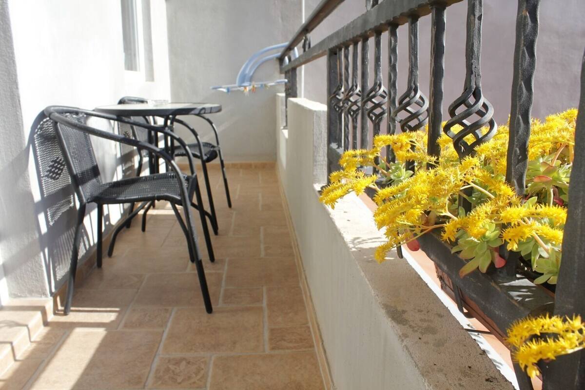 Un balcone con tavolo e sedie, una ringhiera adornata da fiori gialli e vista sulla strada.