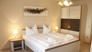 Una cama bien hecha con sábanas y toallas blancas, una lámpara de noche, una mesita auxiliar pequeña, un espejo grande y una obra de arte colgada en la pared.